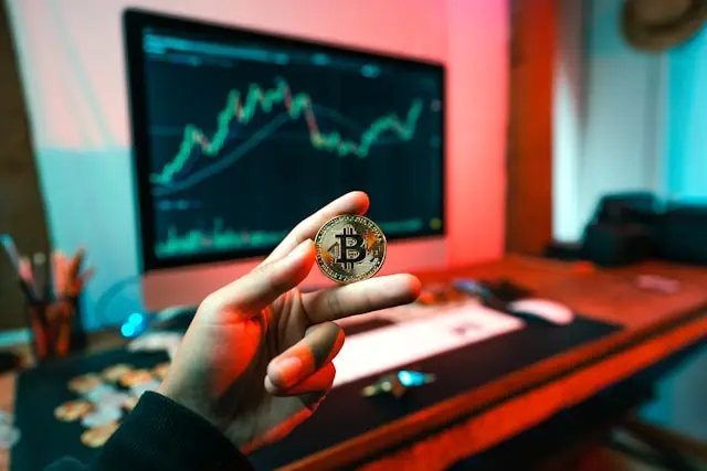 Hand hält eine Bitcoin-Münze vor einem Monitor mit steigendem Kerzenchart als Bezug zum Börsengang der Krypto-Börse Gemini an der Nasdaq.