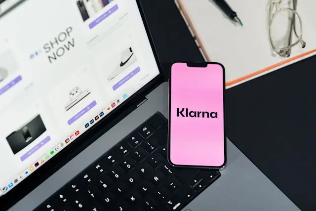 Klarna-Logo auf einem Smartphone-Display neben einem Laptop mit Onlineshop im Zusammenhang mit erwarteten Kursausschlägen
