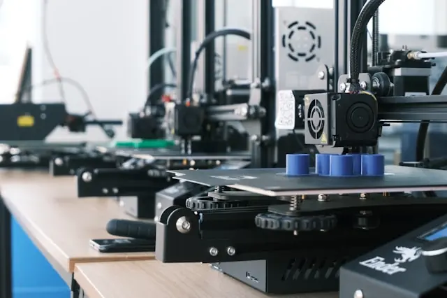 Mehrere 3D-Drucker in einer Werkstatt oder einem Labor drucken blaue Kunststoffobjekte auf Druckplatten. Die Maschinen stehen nebeneinander auf einem Arbeitstisch in moderner Umgebung.