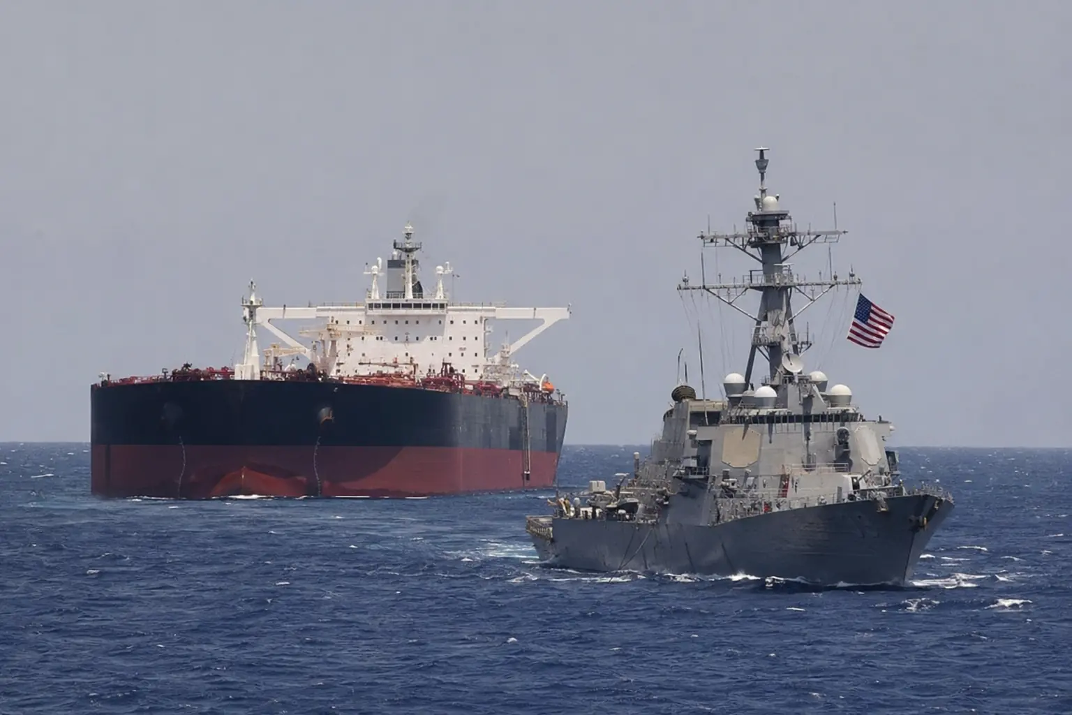 Bildtitel: Ölpreis über 83 Dollar – Öltanker eskortiert von US-Kriegsschiff Großer Öltanker neben einem grauen US-Kriegsschiff mit gehisster amerikanischer Flagge auf offener See als Bezug zur Golf-Eskalation und steigendem Ölpreis