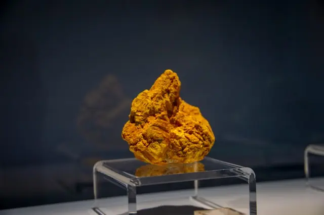 Uranpreis bei 87 Dollar mit Uranerz in einer Museumsvitrine Gelb-oranges Uranerz auf einem Acrylsockel in einer Vitrine vor dem Hintergrund von 87 Dollar je Pfund Uran und knapper Minenförderung
