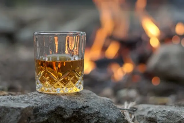 Whisky-Fässer ohne Regulierung mit gefülltem Glas am Lagerfeuer Bernsteinfarbener Whisky in einem Kristallglas auf einem Stein vor offenem Feuer als Bezug zum unregulierten Fassmarkt